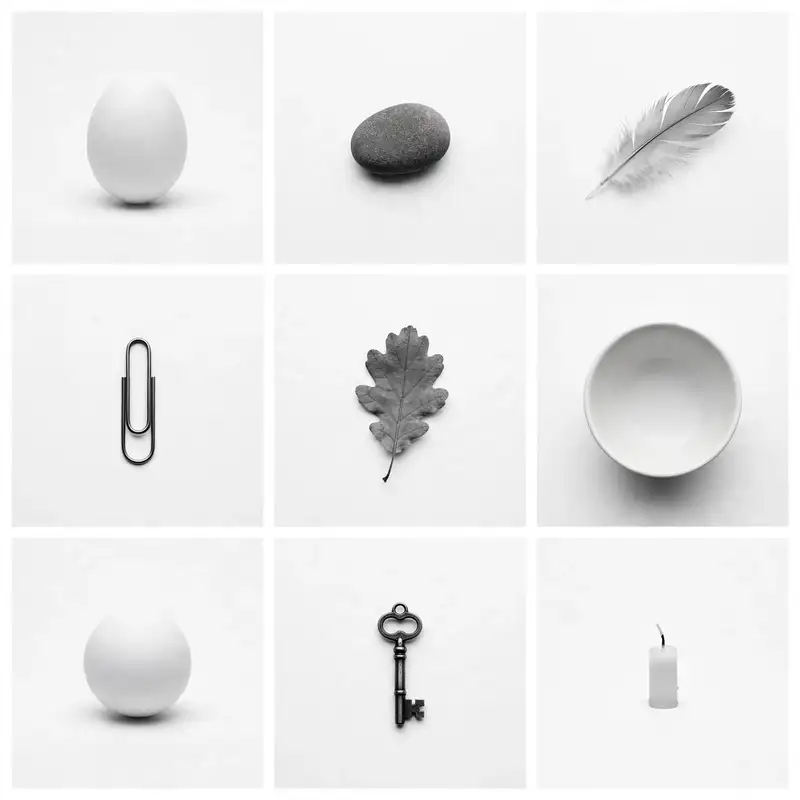 Nine minimal black and white photographs in a 3x3 grid: a white egg, a smooth pebble, a feather, a paperclip, an oak leaf, an empty bowl, a white sphere, a small antique key, a candle
